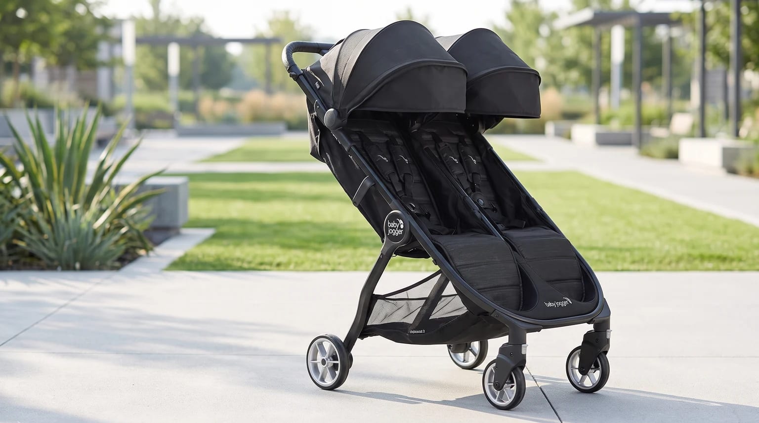 The black Baby Jogger side-by-side double buggy standing empty on a pavement in a sunlit, landscaped park.