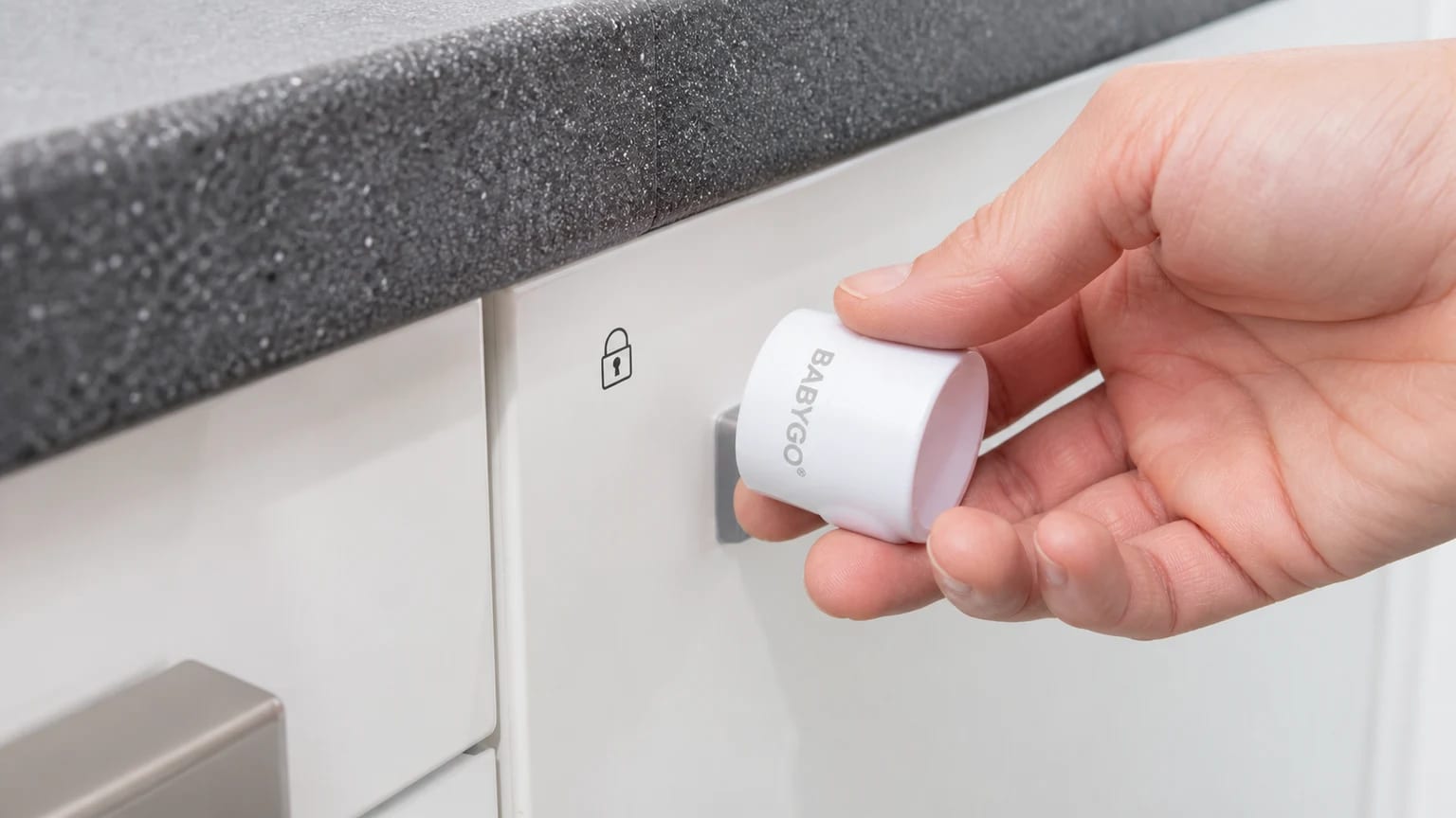 A hand holds a white BABYGO magnetic key against a white kitchen cupboard door, beside a small padlock marker.