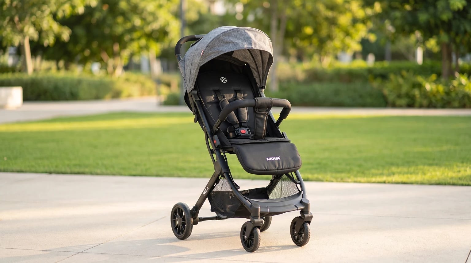 A black pushchair with a grey marl fabric hood, standing on a paved path in a park on a sunny day.