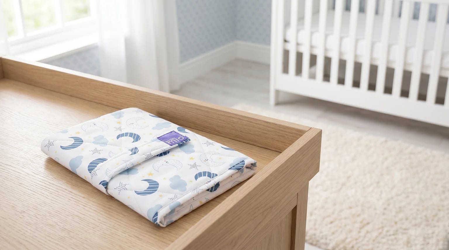 A folded portable changing mat with a blue and yellow moon and stars pattern, sitting on a light oak changing table in a nursery.