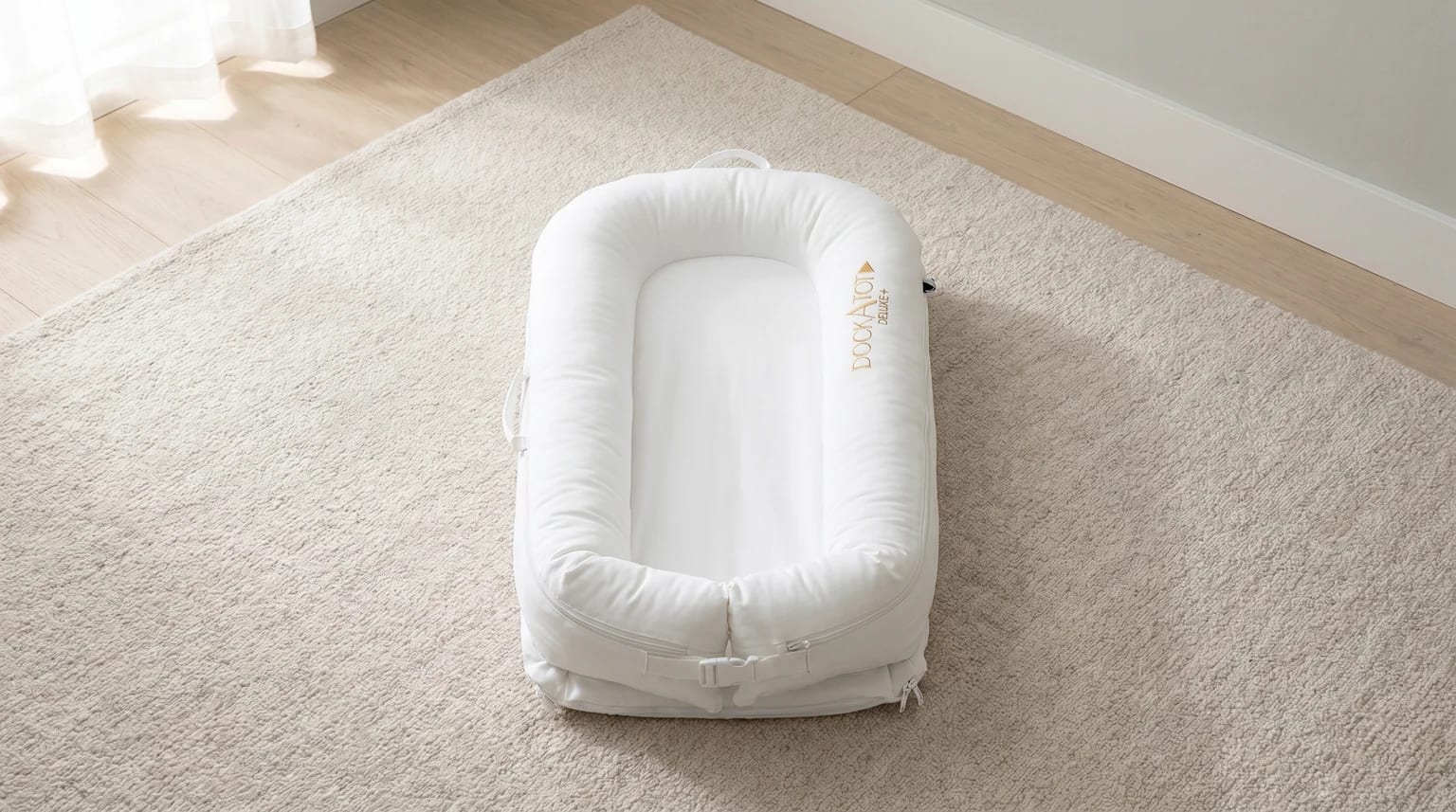 A white DockATot Deluxe+ baby pod placed on a soft, light-coloured rug in a brightly lit room.