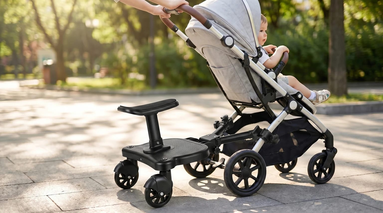 A black buggy board with a seat attached to a grey pushchair being pushed by a parent along a pavement in a park.