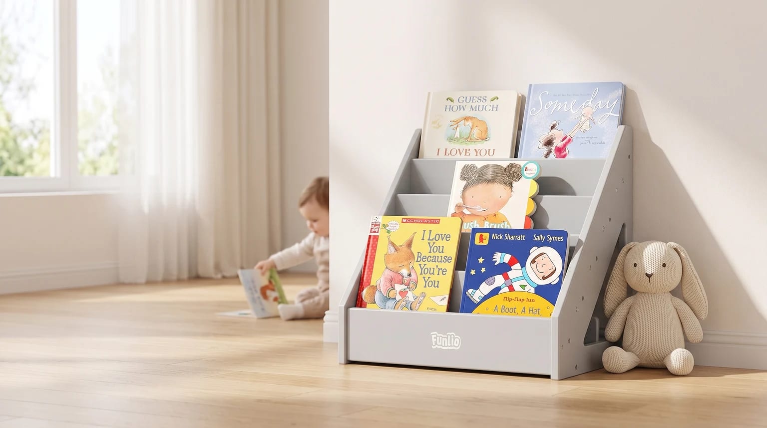 A grey, front-facing children's bookcase on a wooden floor, filled with picture books. A knitted rabbit toy sits beside it and a baby reads in the background.