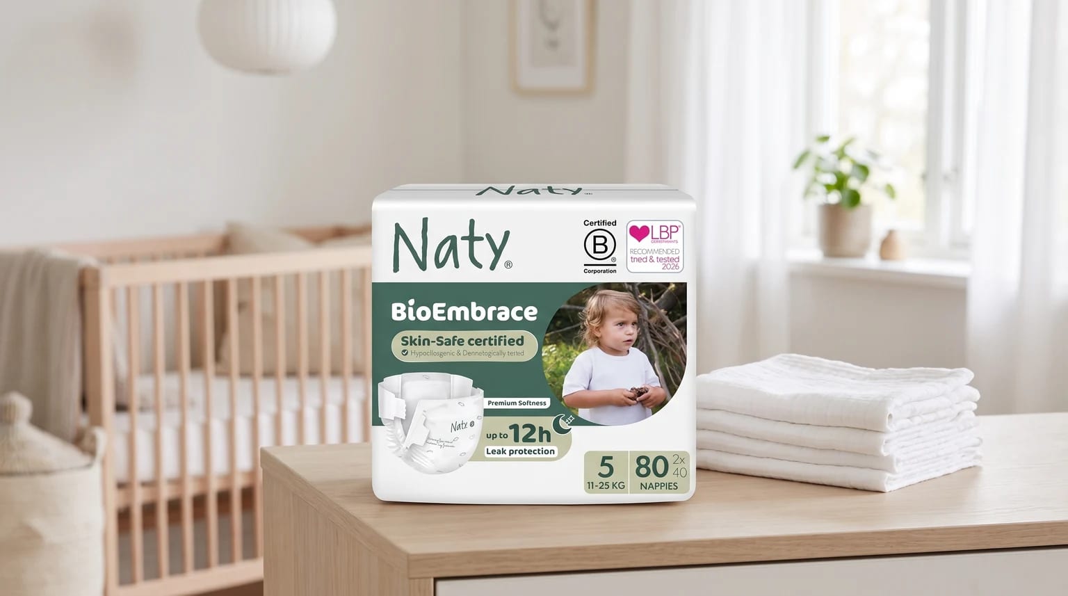 A pack of Naty BioEmbrace nappies sits on a wooden dresser in a nursery, next to a stack of white muslin cloths, with a baby's cot in the background.