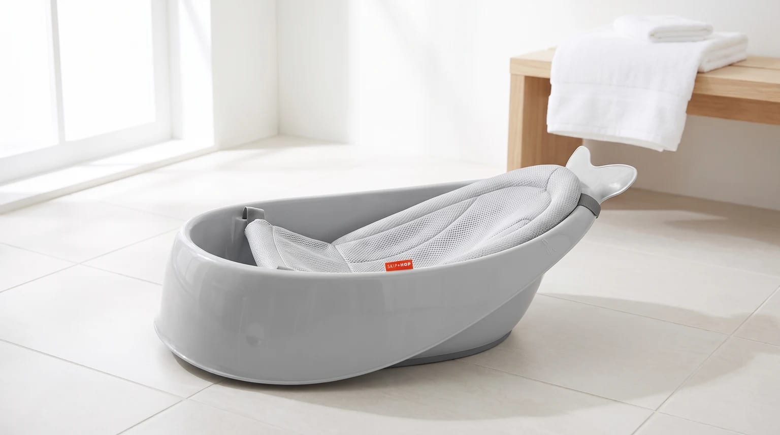 A grey, whale-shaped baby bath with a mesh sling insert, sitting on a brightly lit, white-tiled bathroom floor.