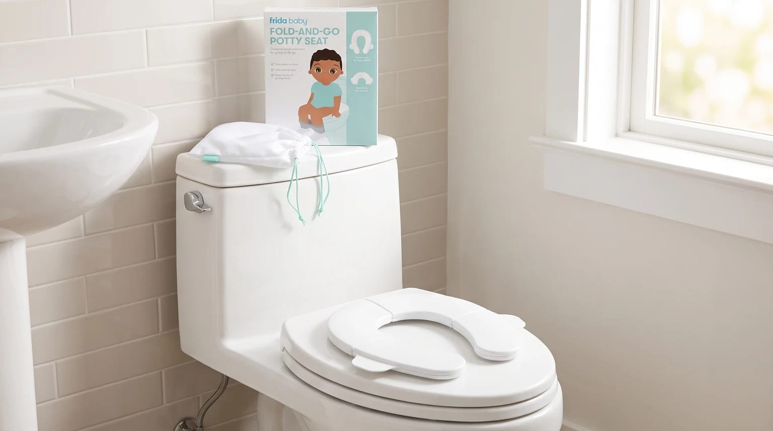 A white foldable travel potty seat placed on a toilet in a bathroom, with its box and drawstring travel bag resting on the cistern.