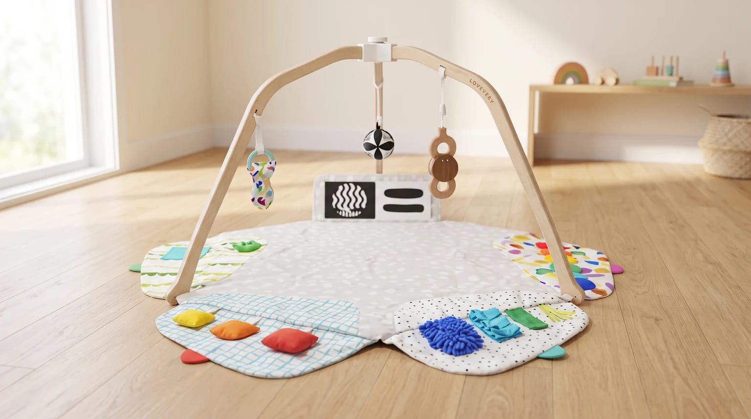 A wooden-framed baby play gym with a sensory mat laid out on a light wood floor. Three different toys hang from the arch over the colourful mat.