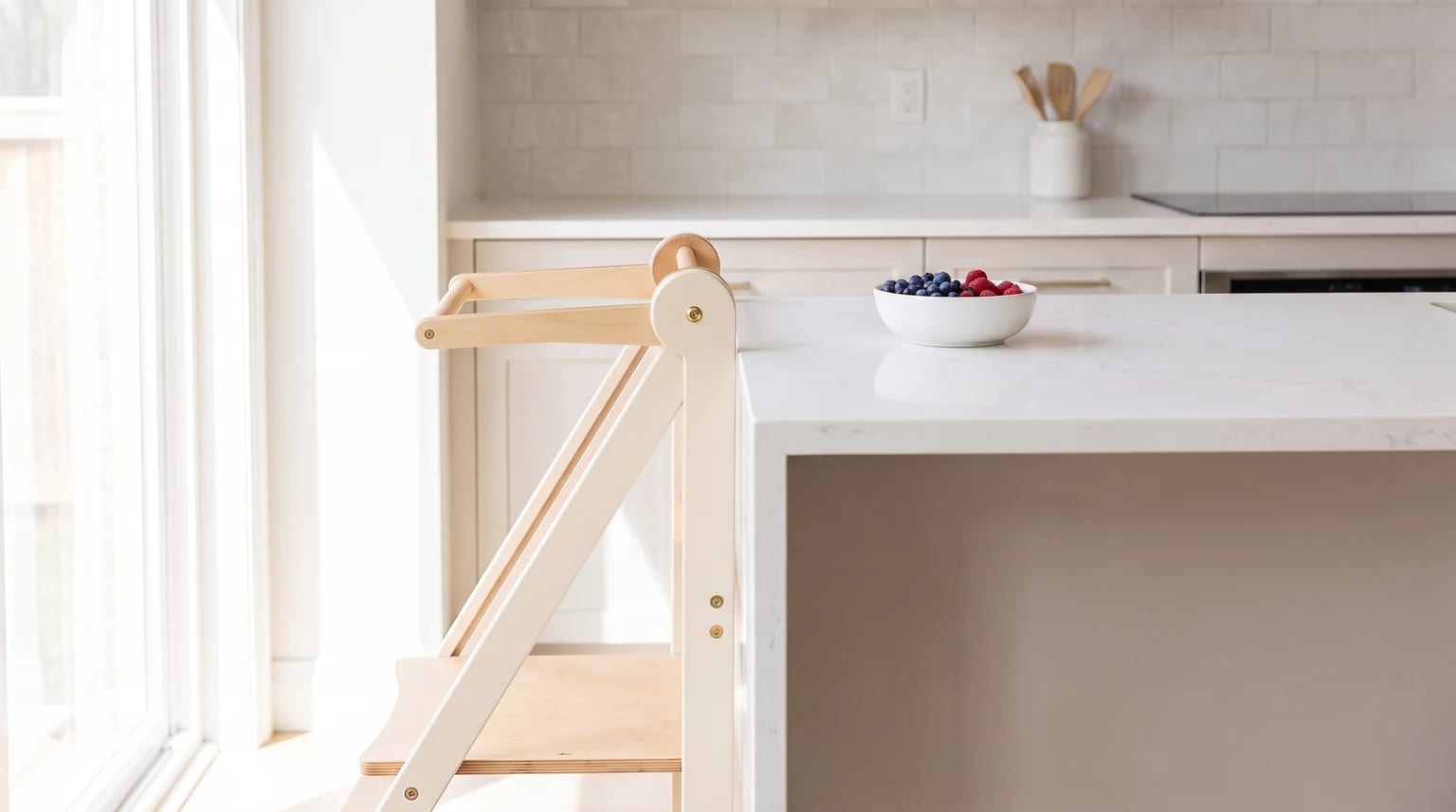 A wooden learning tower with a white finish, standing by a kitchen island with a bowl of fresh berries on the white worktop.