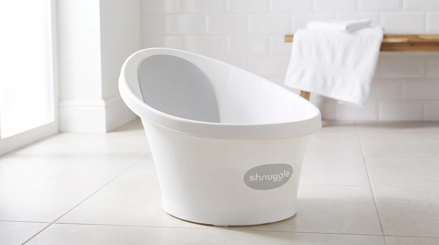 The white Shnuggle baby bath with its grey backrest, standing on a tiled floor in a bright, modern bathroom.