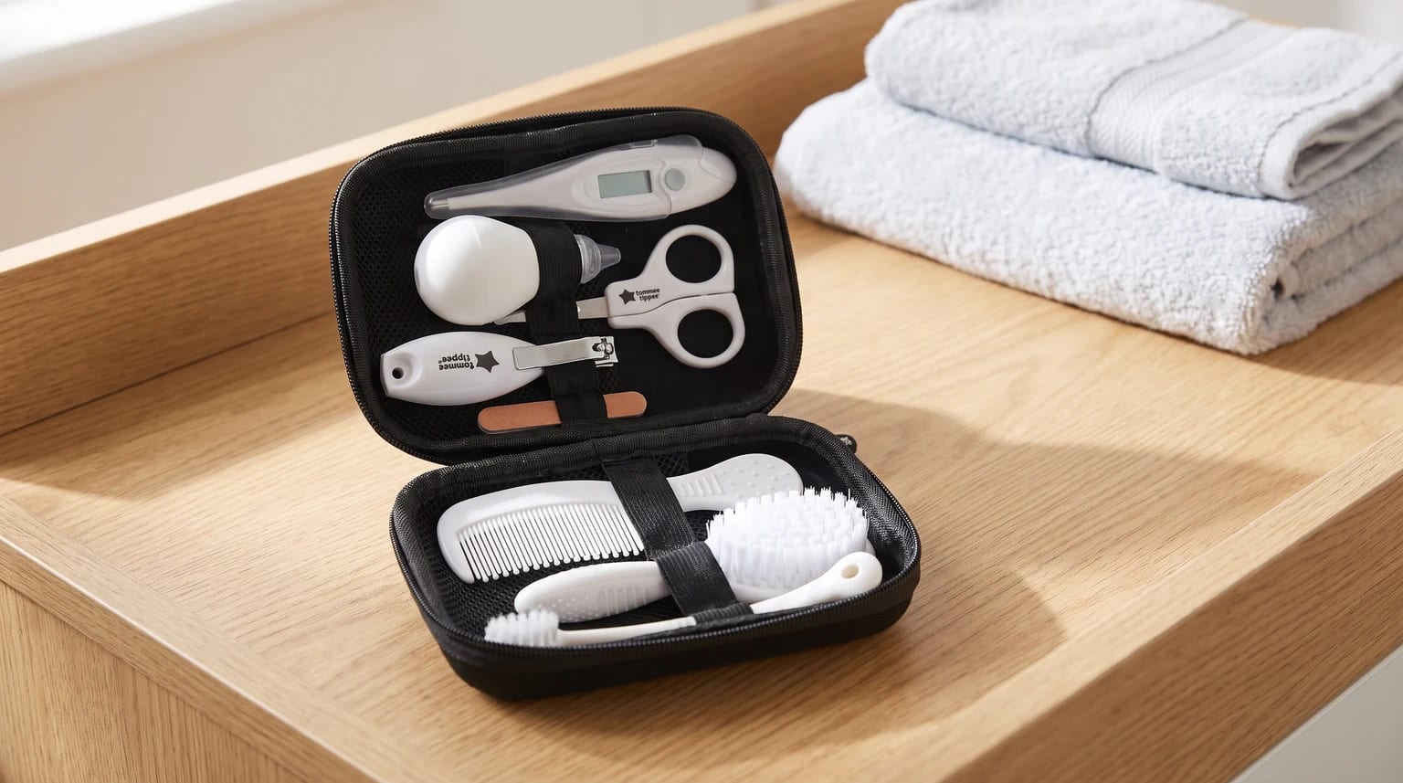 An open black travel case displaying a baby healthcare and grooming set, including a brush, comb, and thermometer, set on a wooden changing unit.