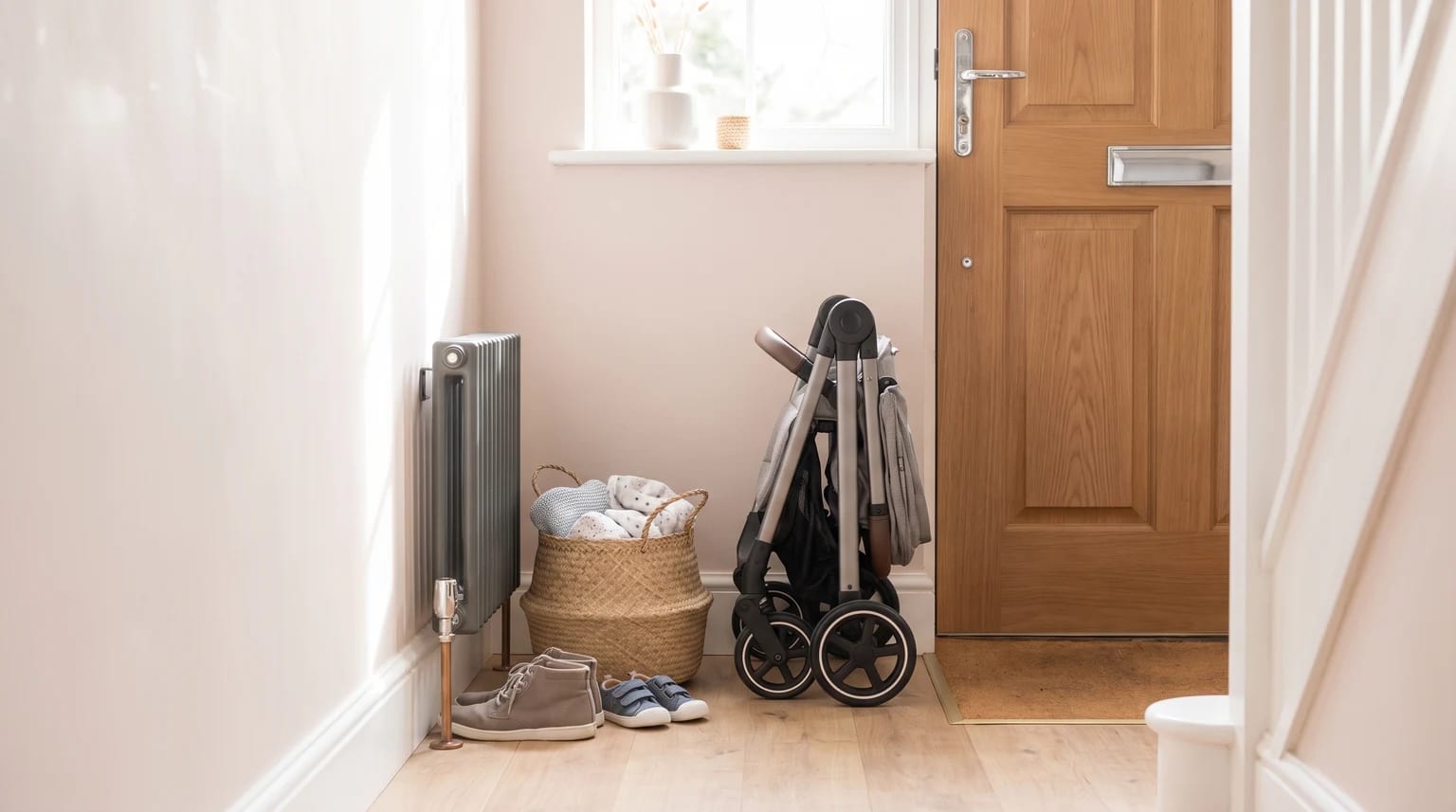 A folded pushchair stands in a sunlit hallway by the front door, with a basket of blankets and shoes on the wooden floor.