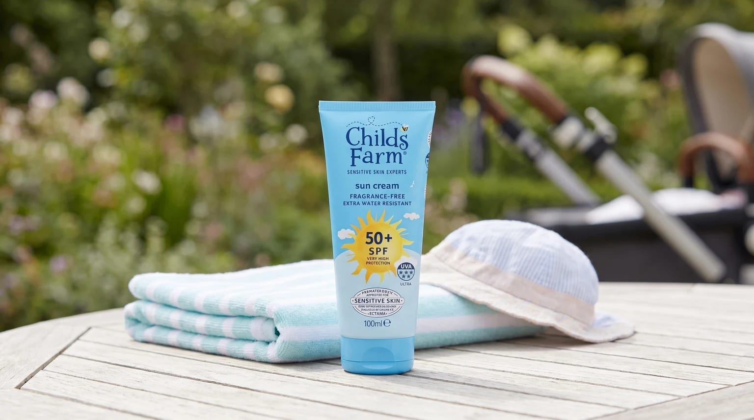 A tube of Childs Farm SPF 50+ sun cream on a wooden garden table, next to a baby's sun hat and a folded towel.