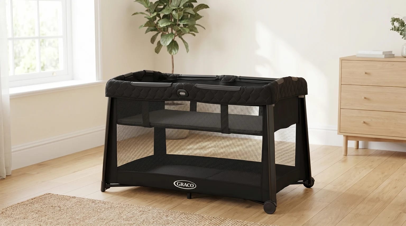 A black Graco FoldLite LX travel cot with bassinet insert set up empty in a light-filled room.