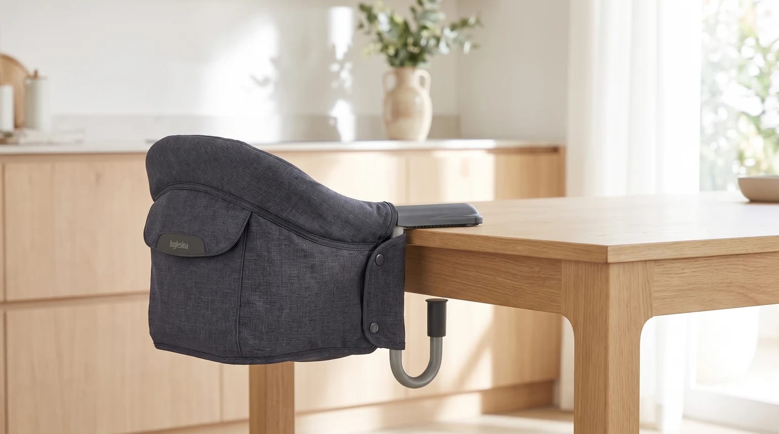 A padded, dark grey fabric hook-on high chair clamped onto the side of a wooden dining table in a bright, contemporary kitchen.