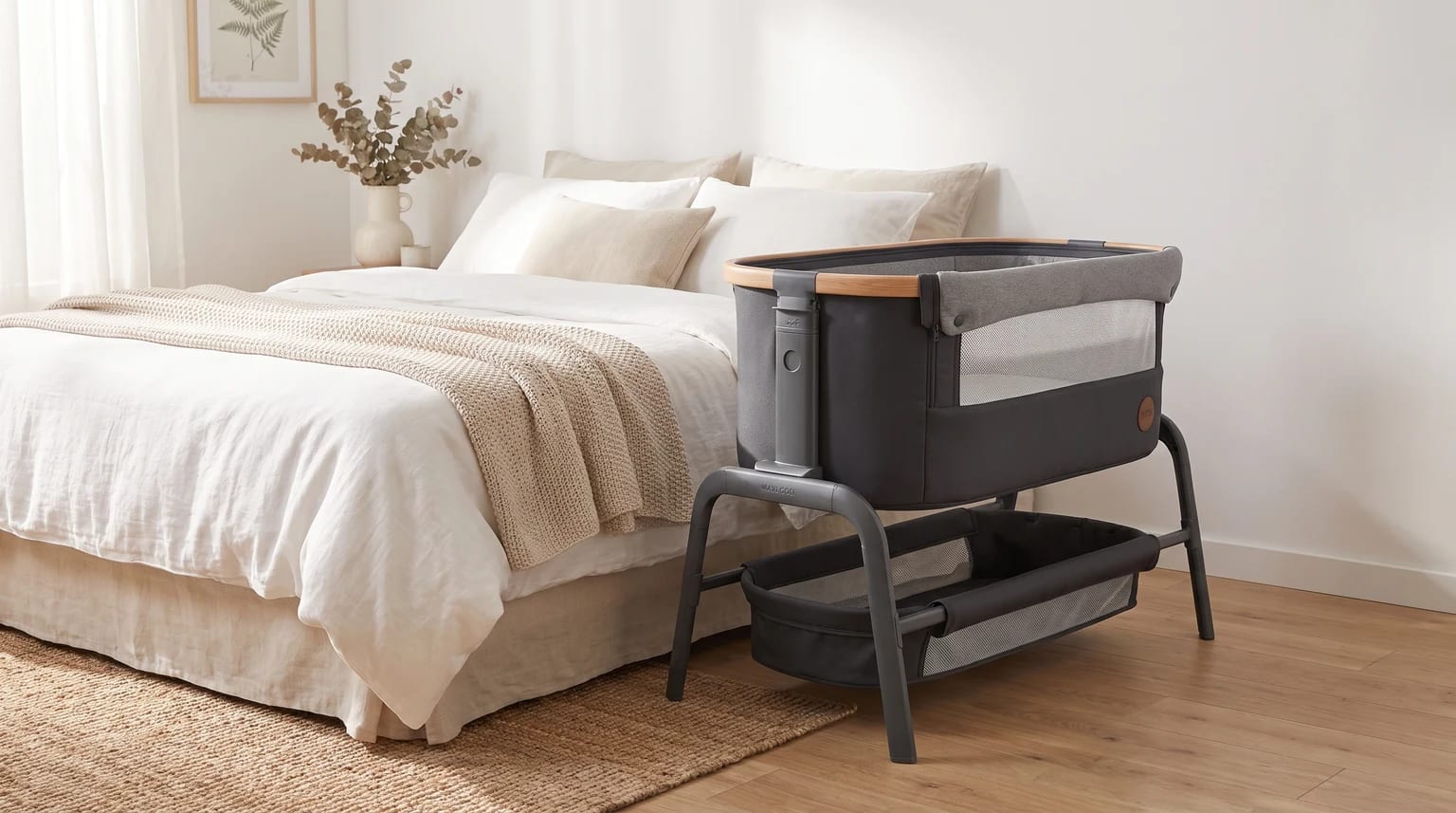 A grey bedside crib with wood-effect trim and a storage shelf, standing next to a double bed with neutral-coloured linen in a bright bedroom.