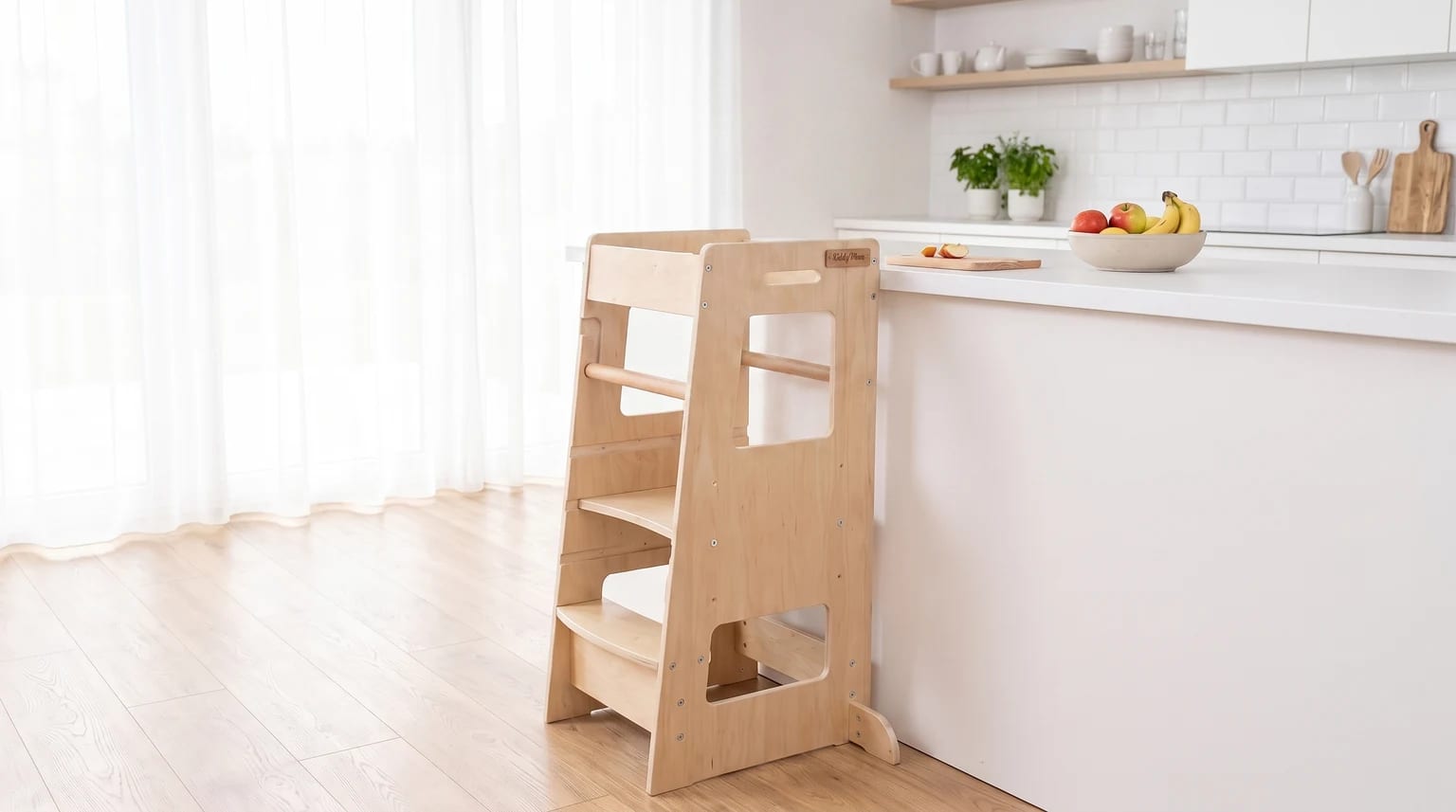A natural wood learning tower stands by a white kitchen worktop with a bowl of fruit and a chopping board.