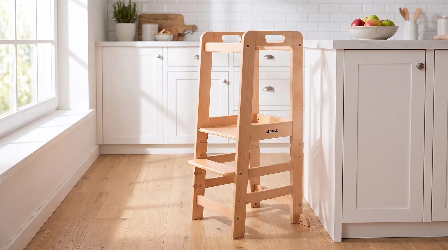 A light wood learning tower for toddlers stands on a wooden floor in a bright, modern kitchen next to white cupboards.
