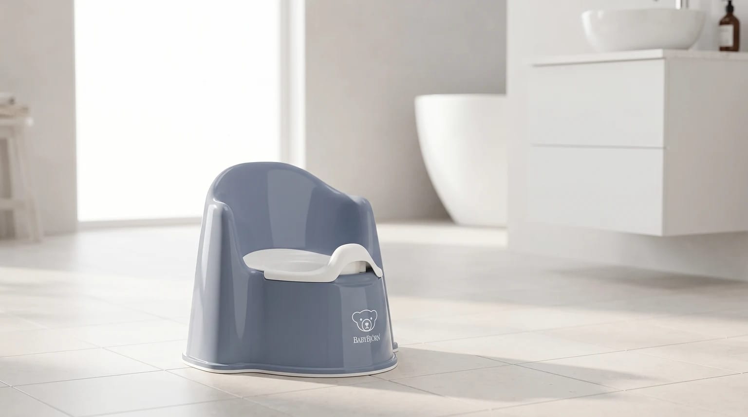 A dusty blue BabyBjörn potty chair with a white inner seat, placed on the tiled floor of a bright, modern bathroom.