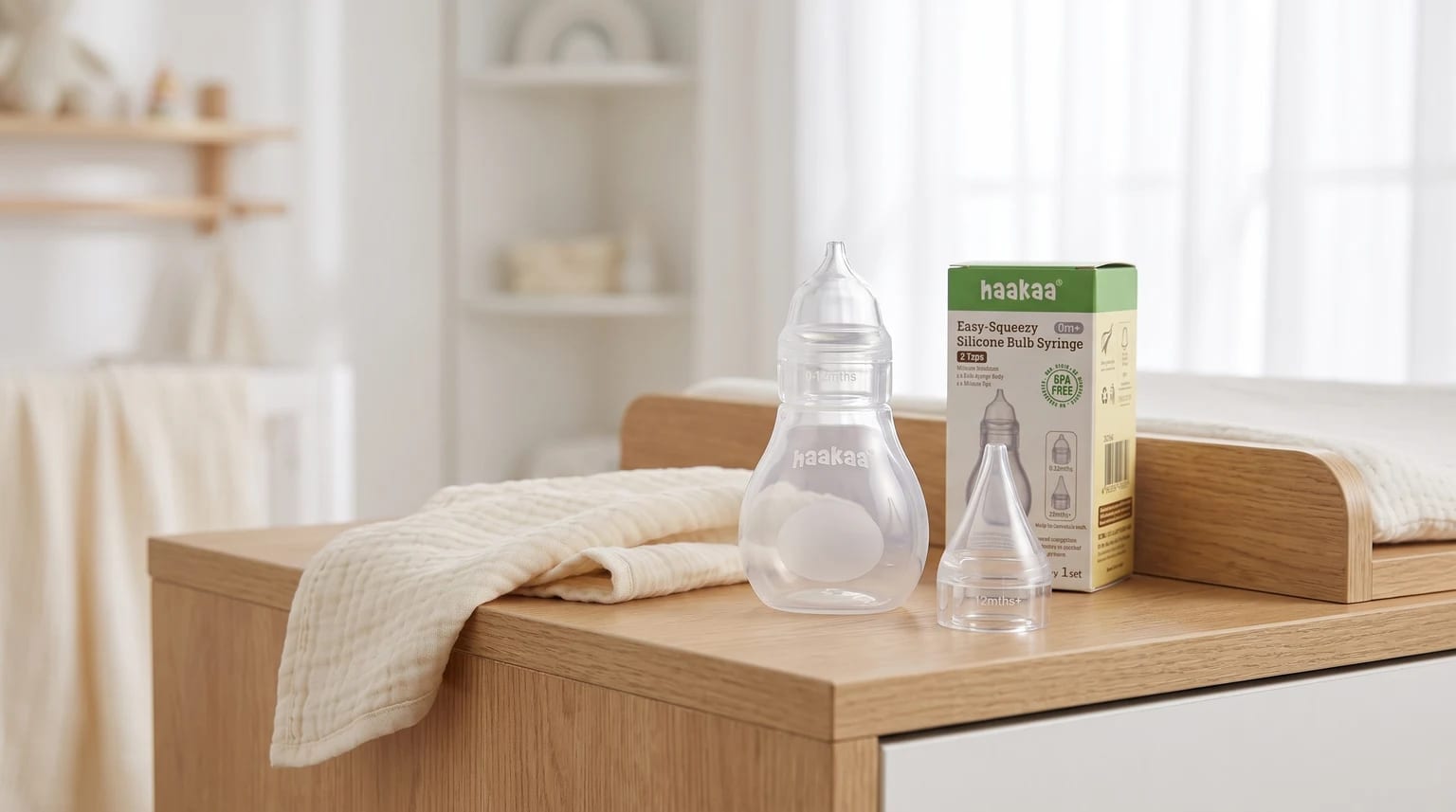A Haakaa silicone bulb syringe and its box sitting on a wooden changing unit next to a cream muslin cloth in a bright nursery.