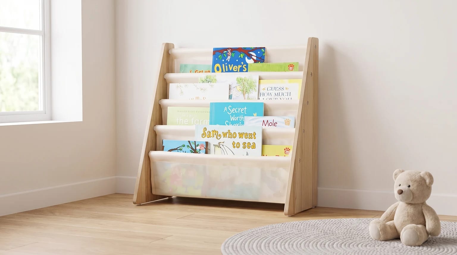 A wooden children's bookcase with cream-coloured fabric slings holding books, placed in the corner of a room with a teddy bear on a rug.