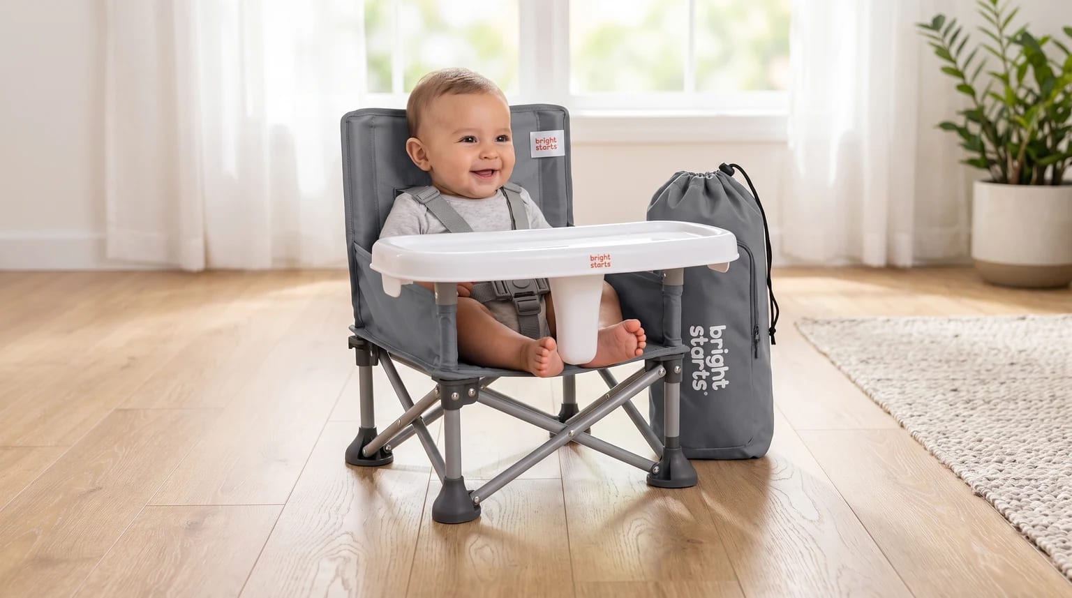 A smiling baby sits in a grey, foldable travel highchair with a white tray, placed on a wooden floor with its carry bag standing beside it.