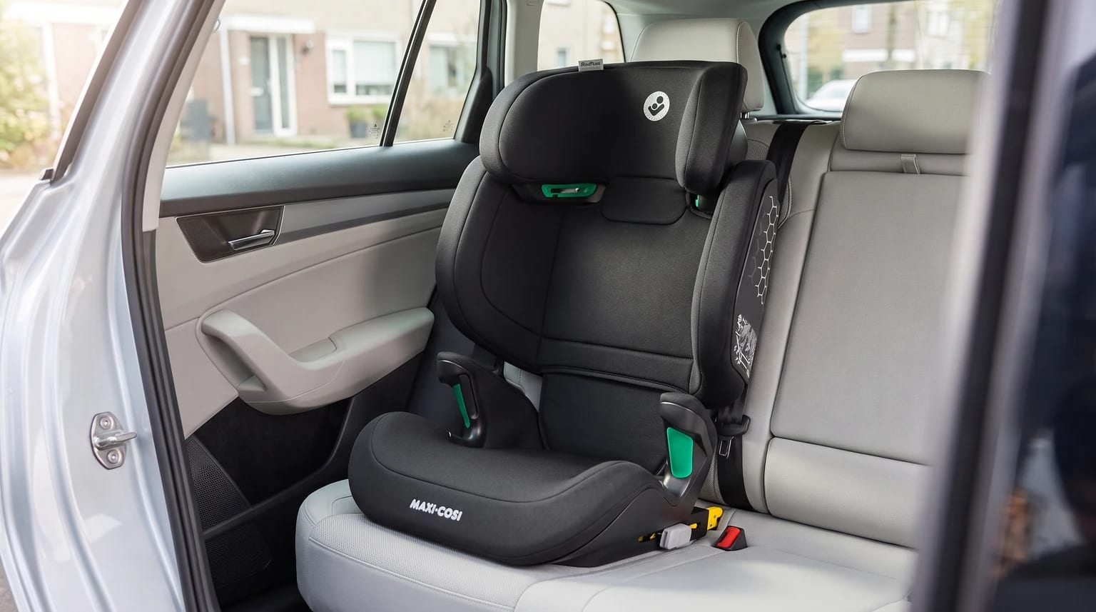 A black Maxi-Cosi high-back booster seat fitted on the light grey back seat of a car, viewed through the open door.