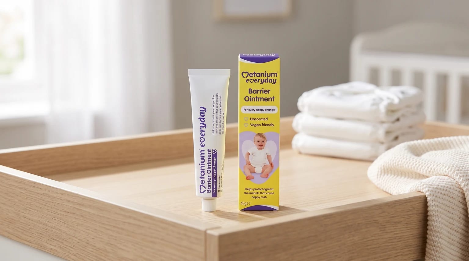 A tube and box of Metanium nappy ointment on a wooden changing unit, with a stack of white nappies and a cot in the background.