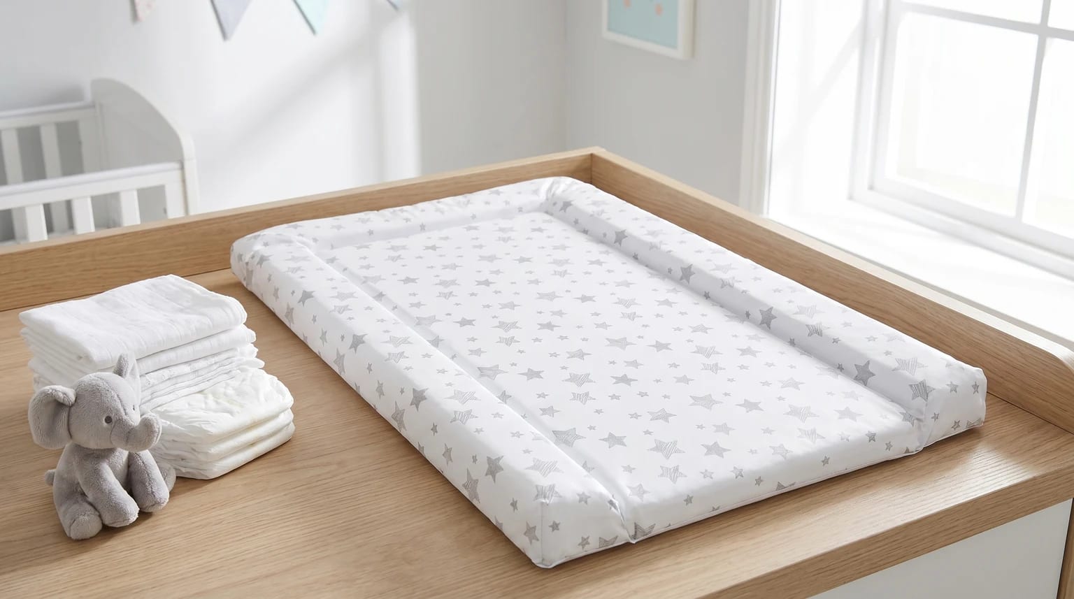 A white changing mat with a grey star pattern, placed on a wooden changing table next to a stack of nappies and a small toy elephant.