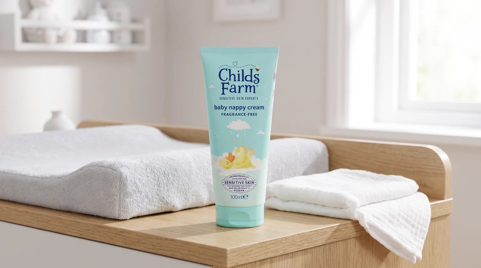 A tube of Childs Farm baby nappy cream on a wooden changing table, next to a soft changing mat and a pile of clean white cloths.