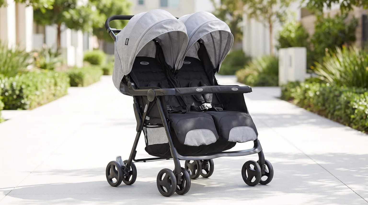 A grey and black side-by-side double pushchair with its hoods up, shown empty on a sunny pavement lined with bushes.