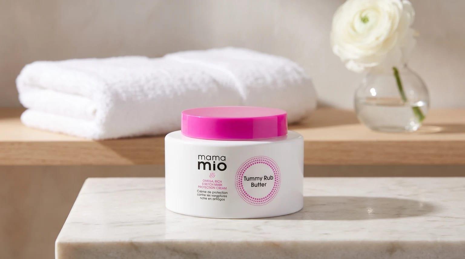 A white tub of Mama Mio Tummy Rub Butter with a bright pink lid, sitting on a marble surface in a calm bathroom setting.