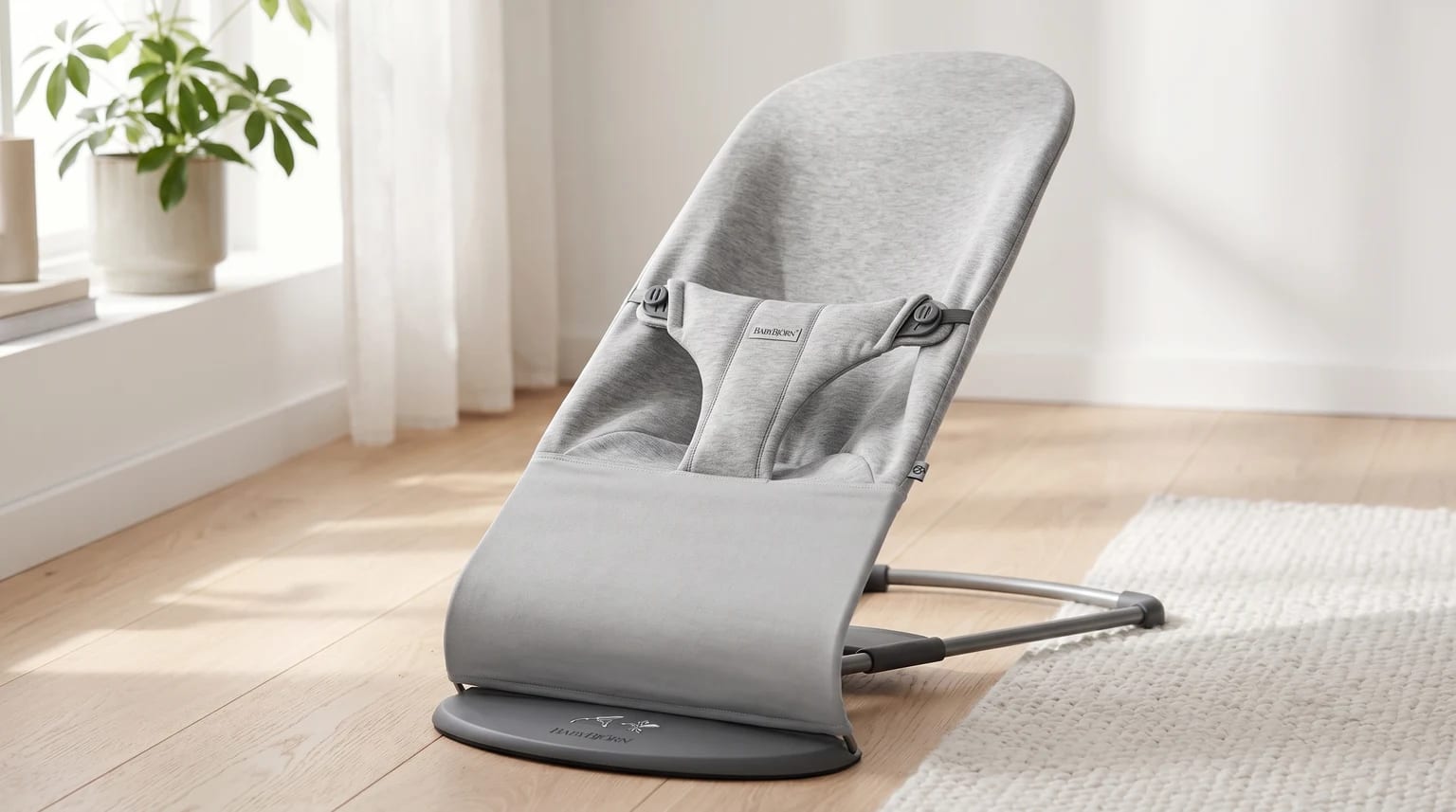 A light grey BabyBjorn baby bouncer with a fabric seat, positioned on a wooden floor in a bright, sunlit room.