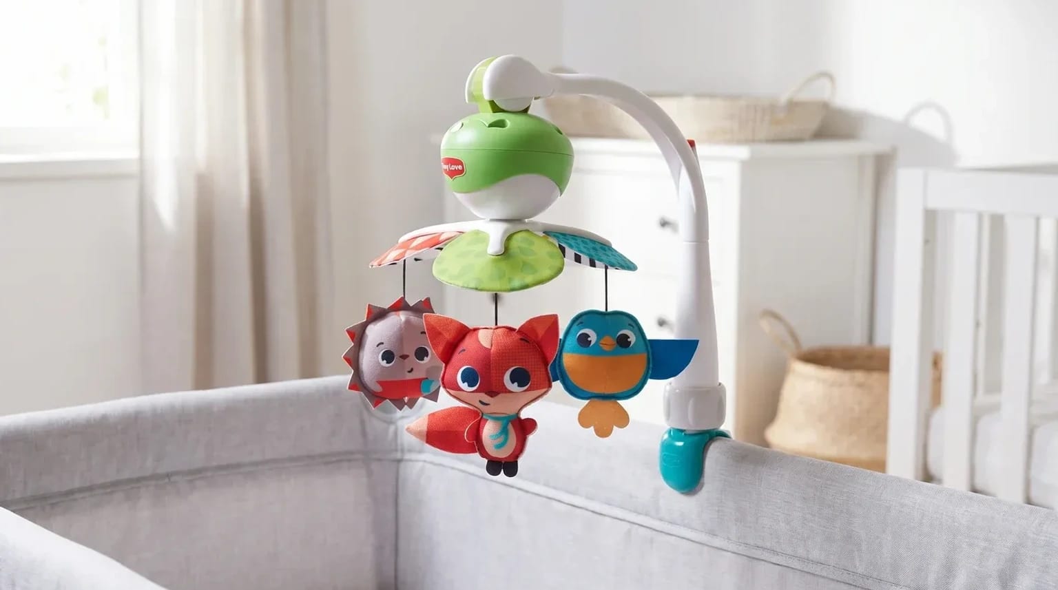 A colourful cot mobile with plush fox, bird, and hedgehog toys, attached to the side of a grey travel cot in a bright nursery.