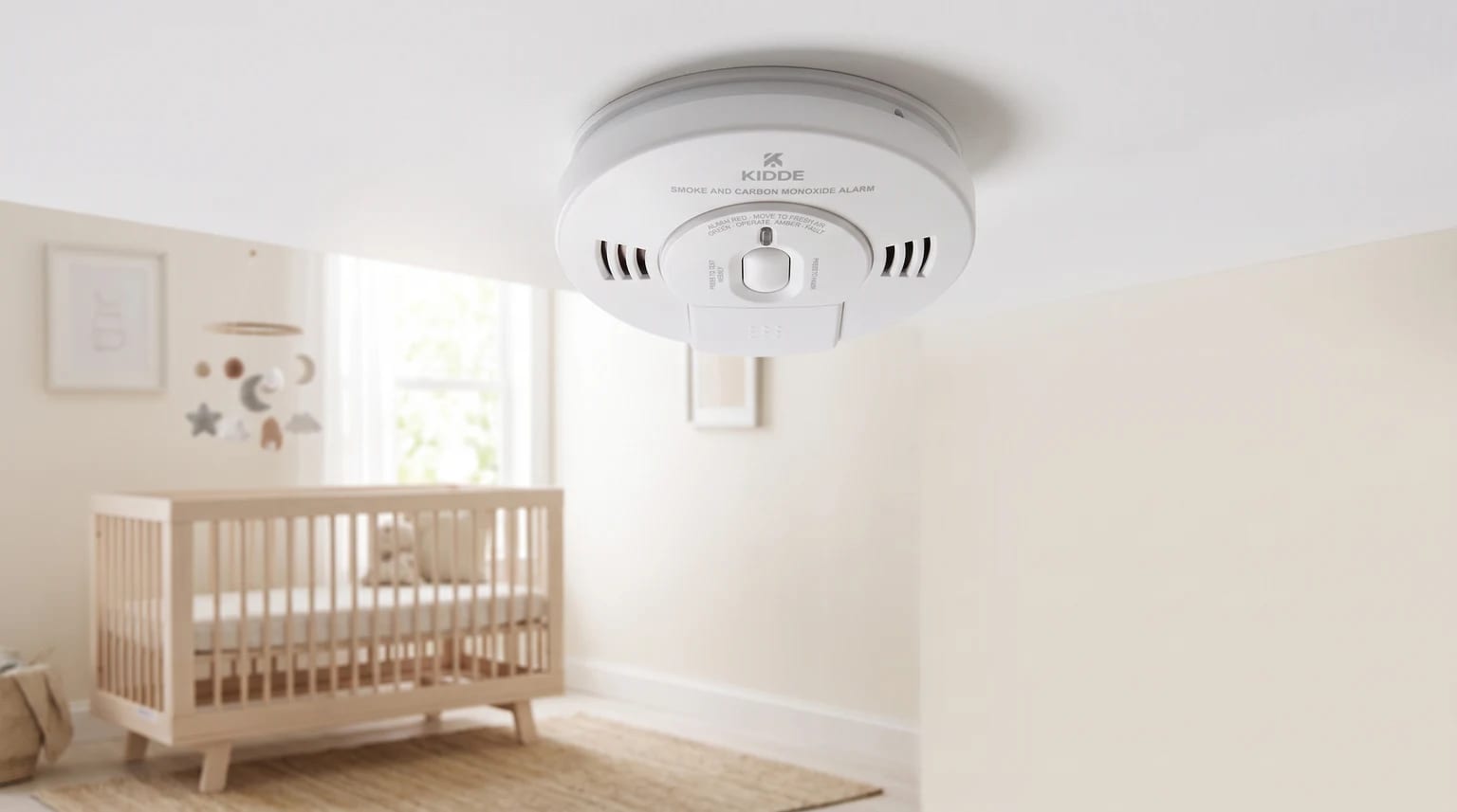 A Kidde smoke and carbon monoxide alarm fitted to the ceiling of a nursery, with a wooden cot visible in the background.