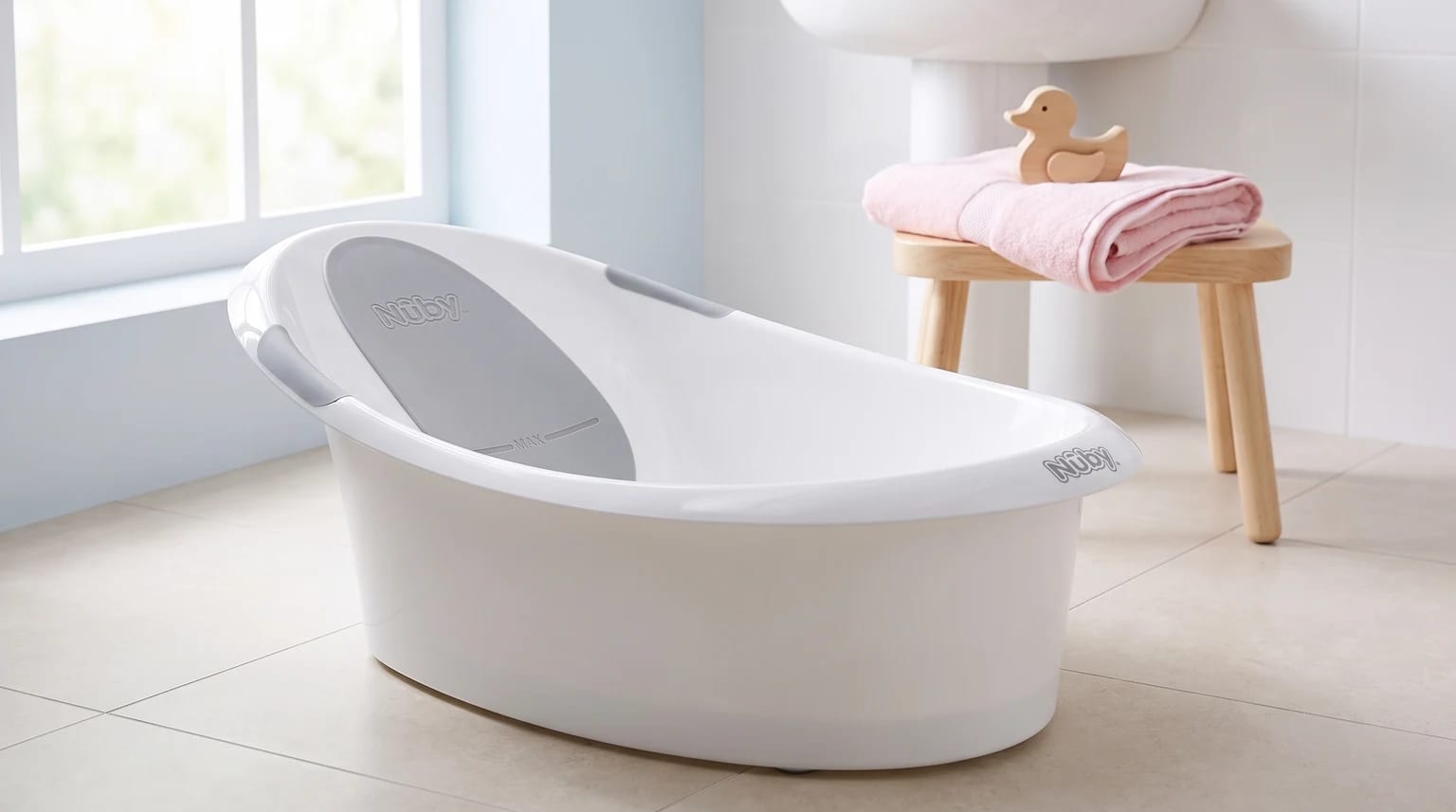 A white and grey Nuby baby bath sits on the floor of a bright bathroom, with a small wooden stool and pink towel in the background.
