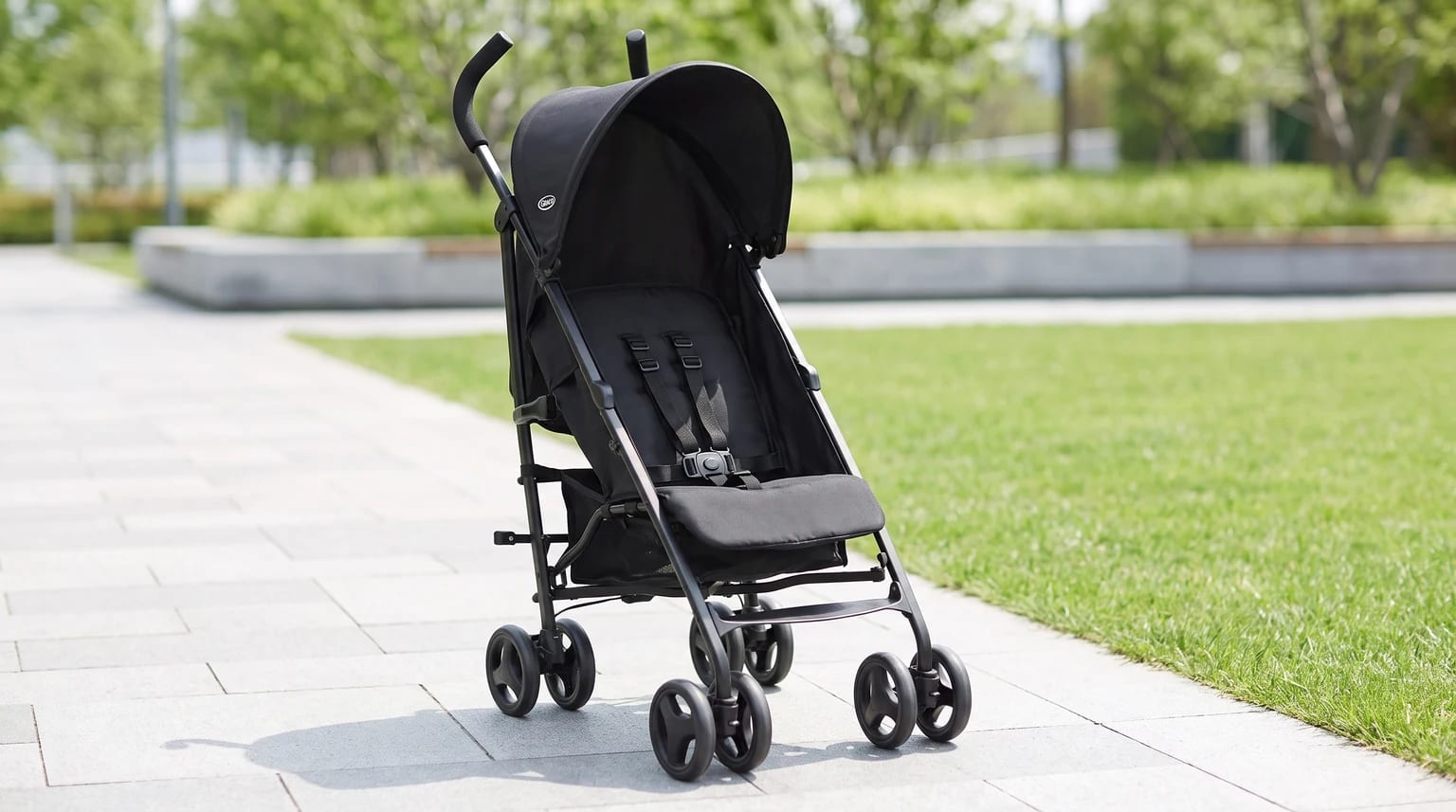 A lightweight black stroller with its sun canopy up, standing empty on a paved path next to a lawn in a park.