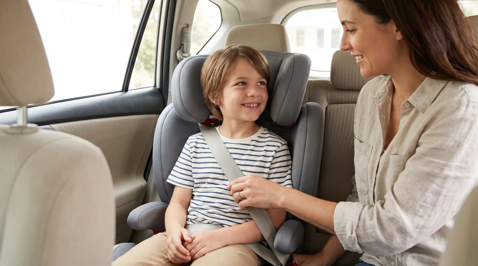 A woman in the back of a car smiles at a young boy as she adjusts the seatbelt on his grey high-back booster seat.