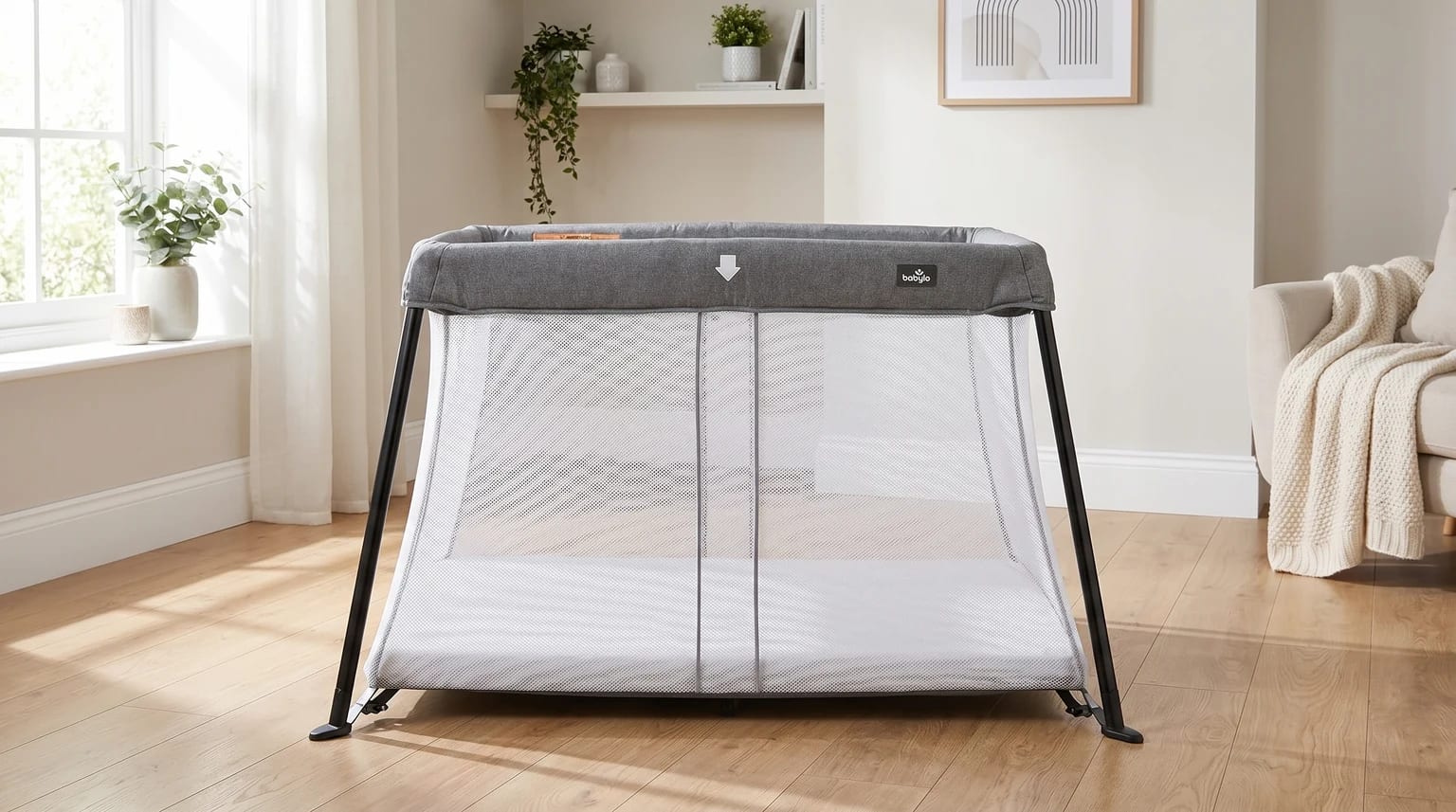 A grey Babylo Liteway travel cot with white mesh sides set up empty in a bright, modern room.