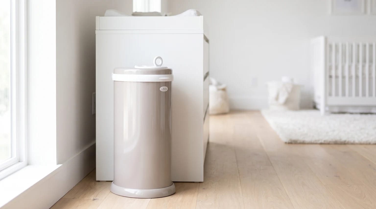 A taupe-coloured Ubbi nappy bin standing on a light wooden floor next to a white changing unit in a bright nursery.