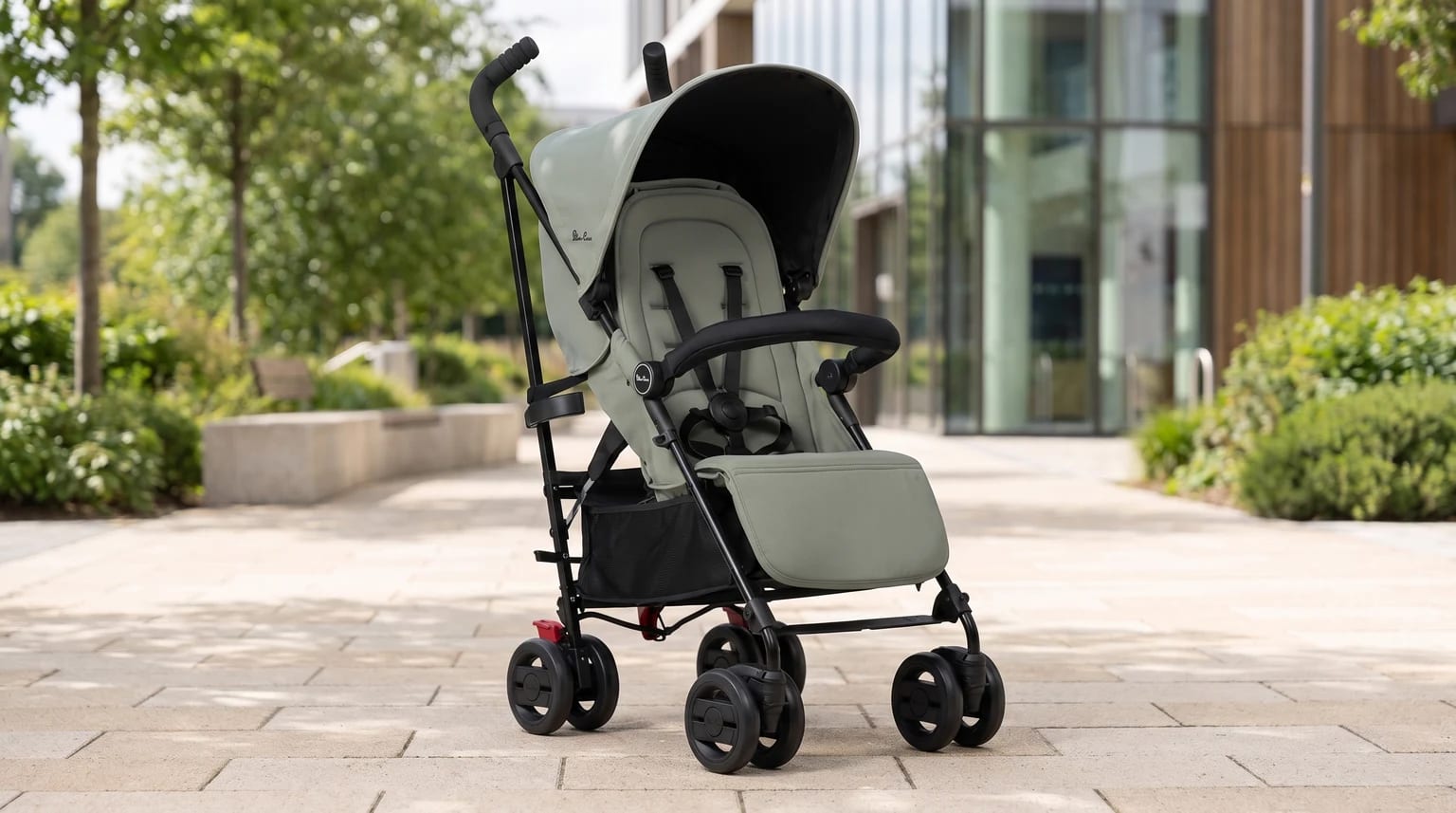 A sage green lightweight pushchair with a black frame, standing empty on a paved walkway in a modern urban setting.