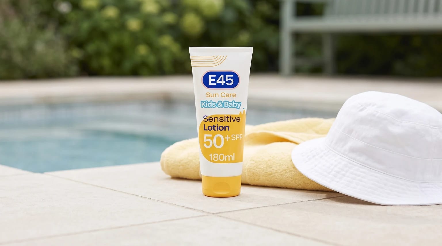 A tube of E45 Kids & Baby sun cream sits beside a swimming pool, next to a yellow towel and a white bucket hat.