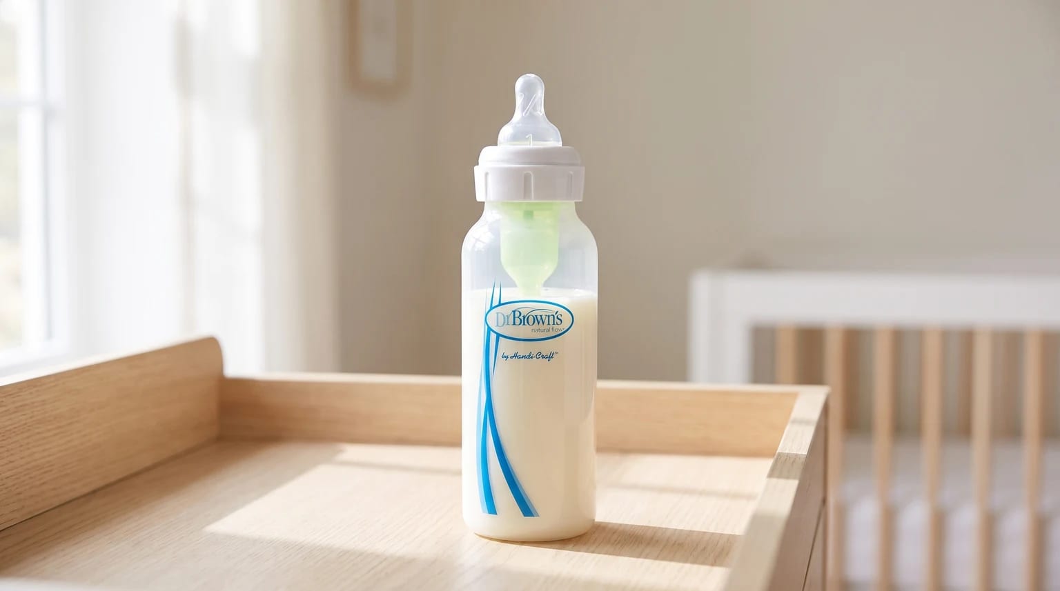 A Dr Brown's baby bottle filled with milk sits on a wooden changing unit in a softly lit nursery, with a cot visible in the background.