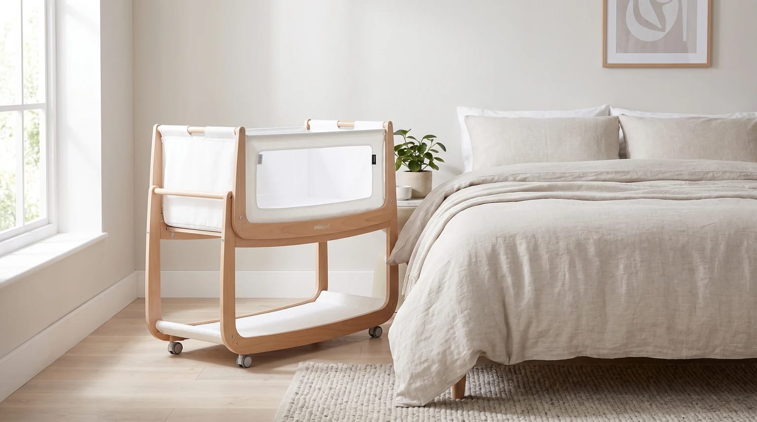 A light wood and white bedside crib positioned next to a parent's bed with neutral linen bedding in a brightly lit bedroom.