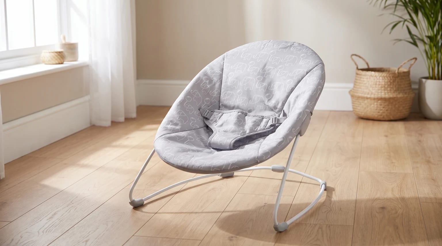 A grey baby bouncer with a white elephant print, sitting on a wooden floor in a sunlit room.
