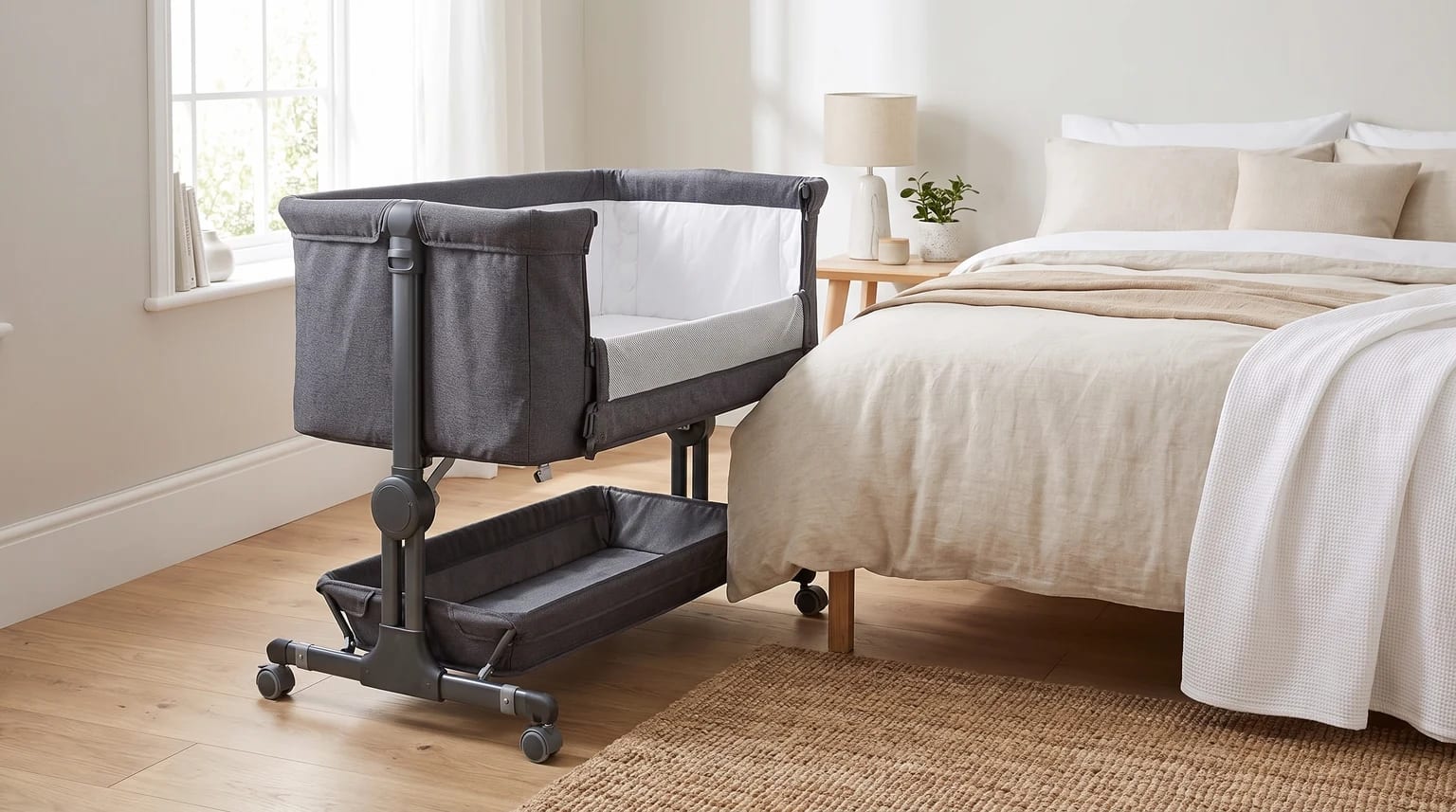A dark grey bedside crib with its side lowered, positioned next to a parent's bed in a light and airy bedroom.