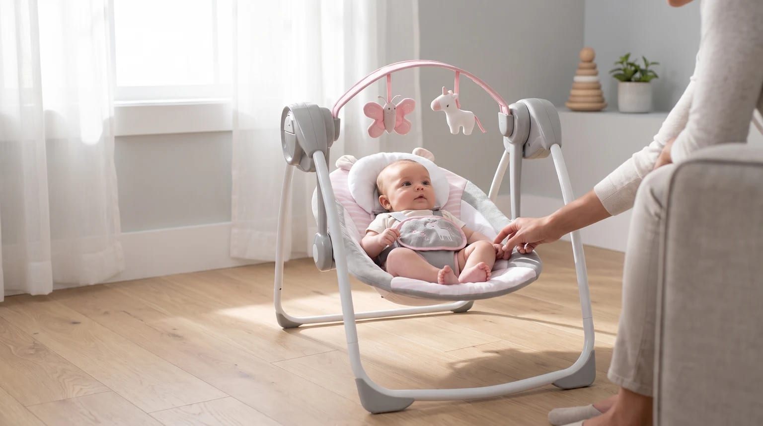 An awake baby sits reclined in a grey and pink baby swing with a toy bar overhead, as a parent reaches out in a bright living room.
