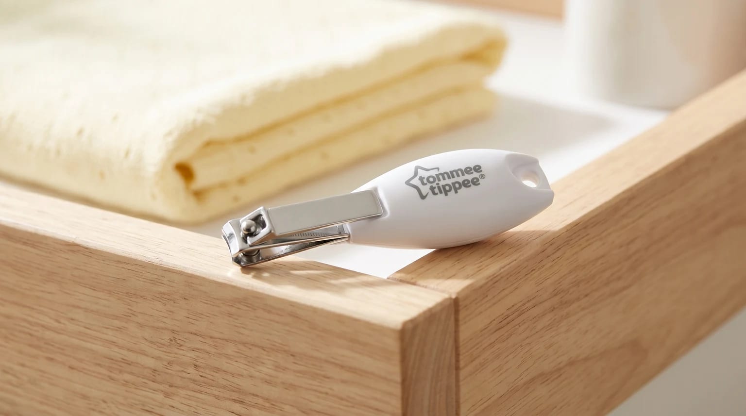 A pair of white Tommee Tippee baby nail clippers resting on the corner of a light wood surface, with a folded yellow towel in the background.