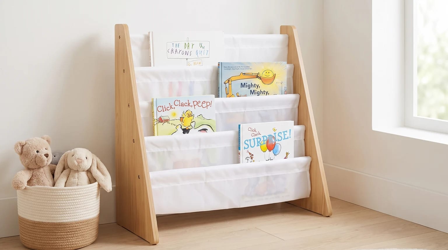 A wooden sling bookshelf filled with children's books, standing on a light wood floor next to a woven basket holding a teddy bear and a bunny.