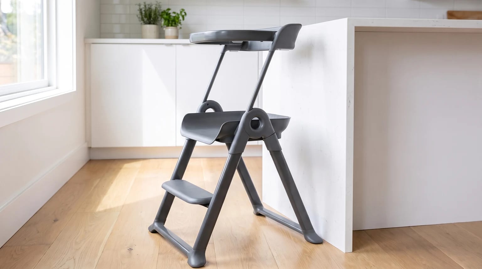 A dark grey toddler learning tower stands on a light wood floor in a modern kitchen, next to a white island.