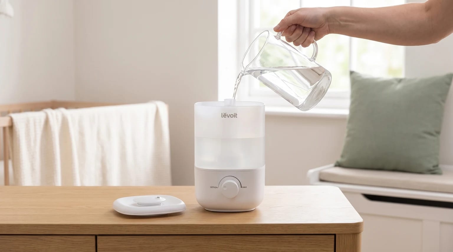 A person pouring water from a glass jug into a white Levoit humidifier, which sits on a wooden table in a nursery.